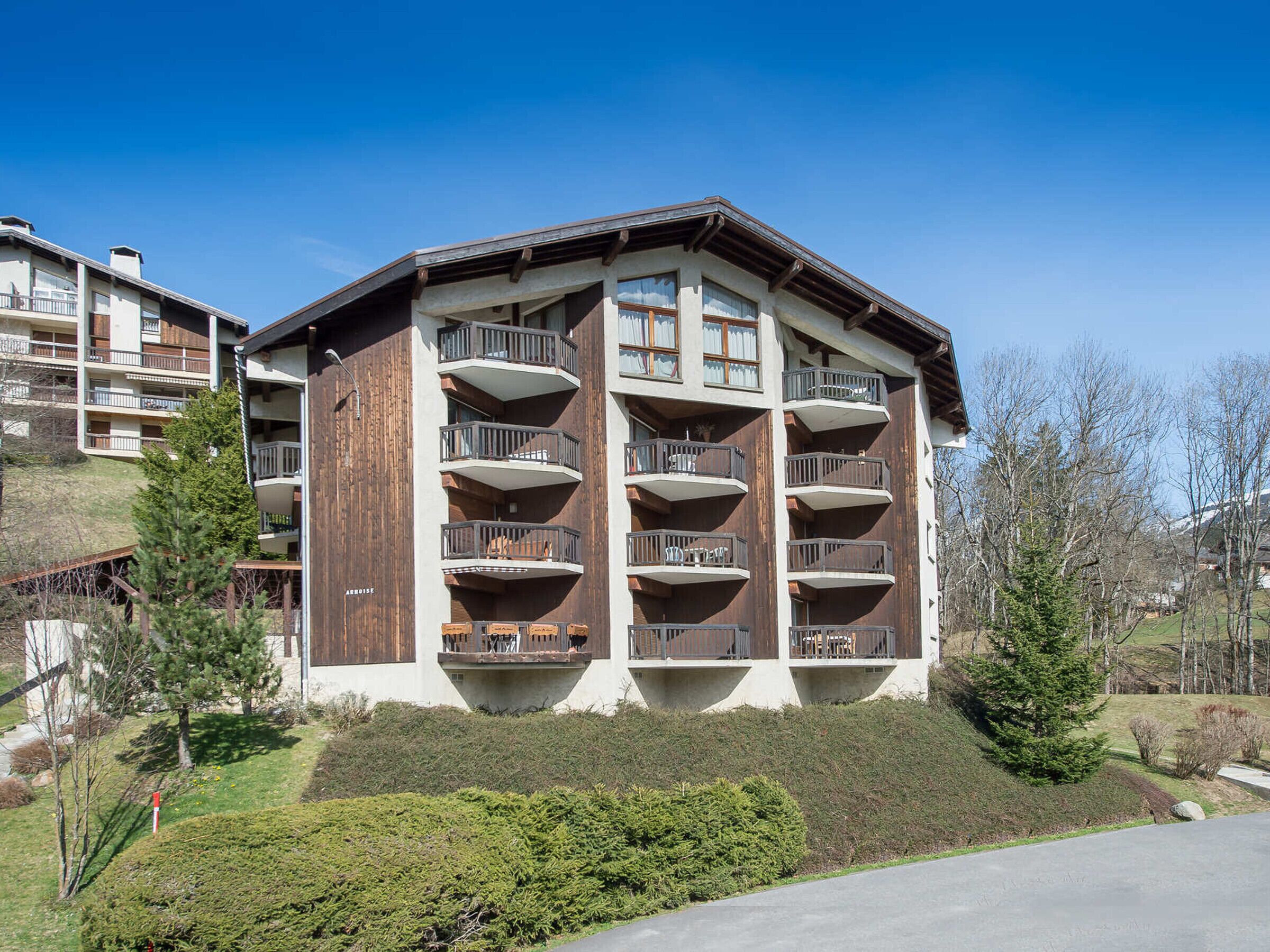 A multi-story building with balconies, likely a hotel or apartment complex, surrounded by trees and a clear sky.