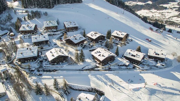 A snow-covered ski resort with multiple chalet-style buildings clustered together, surrounded by trees and a ski slope.