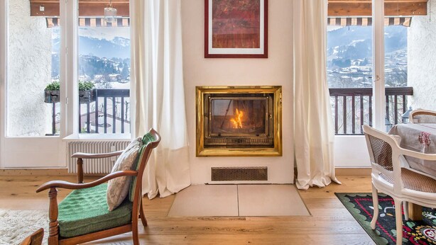 Ein gemütliches Zimmer mit Kamin, Holzboden, einem grünen Sessel und Blick auf eine verschneite Berglandschaft durch große Fenster.