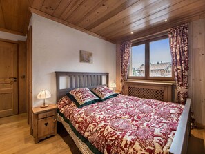 Quarto com teto de madeira, cama com roupa de cama estampada, mesa de cabeceira com abajur e janela com cortinas.
