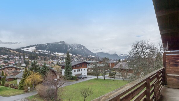 A balcony with a wooden railing overlooking a town with houses, a green lawn, and mountains in the background.