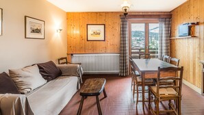A cozy room with a wooden floor, a sofa, a dining table with chairs, a radiator, and a window with a view of a town.
