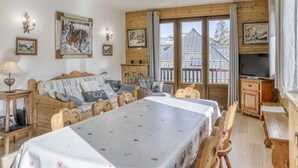 Een gezellige kamer met een houten tafel, een bank en een televisie. Er hangen schilderijen aan de muren en een raam met uitzicht op gebouwen.