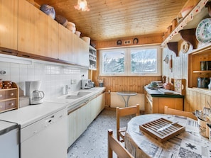 Une cuisine rustique dans une cabane en bois, avec des meubles blancs, une machine à café, un évier, une table à manger avec un set de table, et une fenêtre donnant sur la forêt.