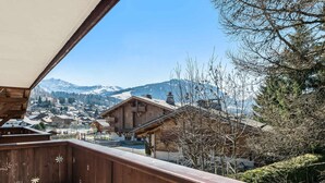 A wooden balcony with a view of a mountain town and snow-capped peaks.