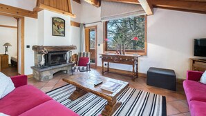 A cozy living room with a stone fireplace, wooden beams, a large window with a view of trees, a red sofa, and a coffee table.