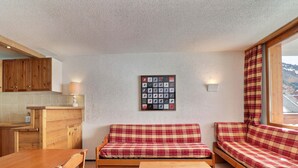 A cozy room with a wooden table, bench, and sofa, a kitchen area, and a wall with a framed picture. - Charming 2-Room, South Balcony, Ski-In/Ski-Out (Méribel)