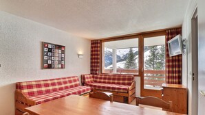 A cozy room with a wooden table, two plaid sofas, a wall-mounted TV, and a window with a mountain view. - Charming 2-Room, South Balcony, Ski-In/Ski-Out (Méribel)