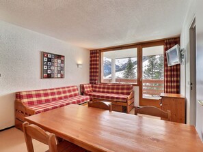 Une pièce chaleureuse avec une table en bois, deux canapés à carreaux, un téléviseur fixé au mur et une fenêtre donnant sur les montagnes.