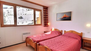 A room with two wooden beds, a window with a view of a snowy landscape, a painting on the wall, and a heater. - Charming 2-Room, South Balcony, Ski-In/Ski-Out (Méribel)