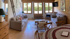 A spacious living room with a sofa, chairs, a coffee table, and a dining table. There are large windows providing natural light and a view of the outdoors.