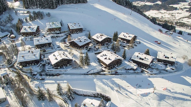 Una località sciistica innevata con diversi edifici in stile chalet raggruppati, circondati da alberi e una pista da sci.