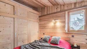 A wooden cabin bedroom with a bed, pillows, a blanket, a window with a view, and a shelf with a candle.