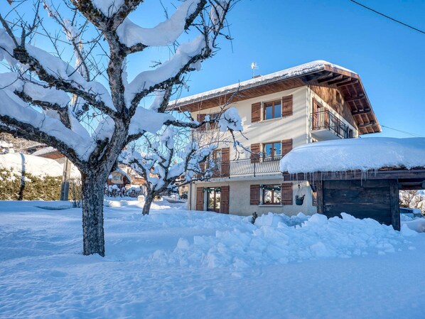 A two-story chalet with a balcony, surrounded by snow-covered trees and a clear blue sky.