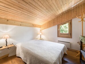 Une chambre avec un plafond en bois, un lit recouvert d’une couette blanche, deux lampes de chevet, une fenêtre donnant sur la verdure et un radiateur.