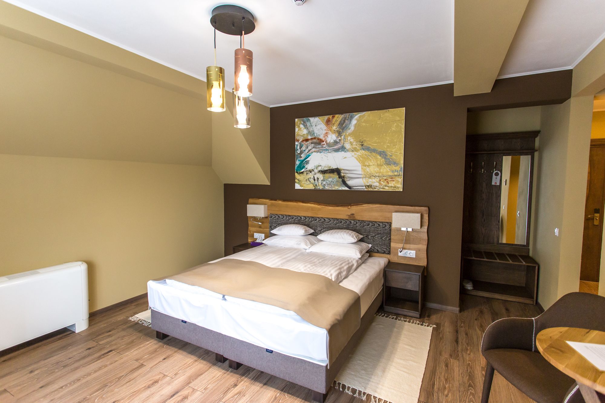 A modern hotel room with a large bed, a wooden headboard, a bedside table, a chair, and a painting on the wall.