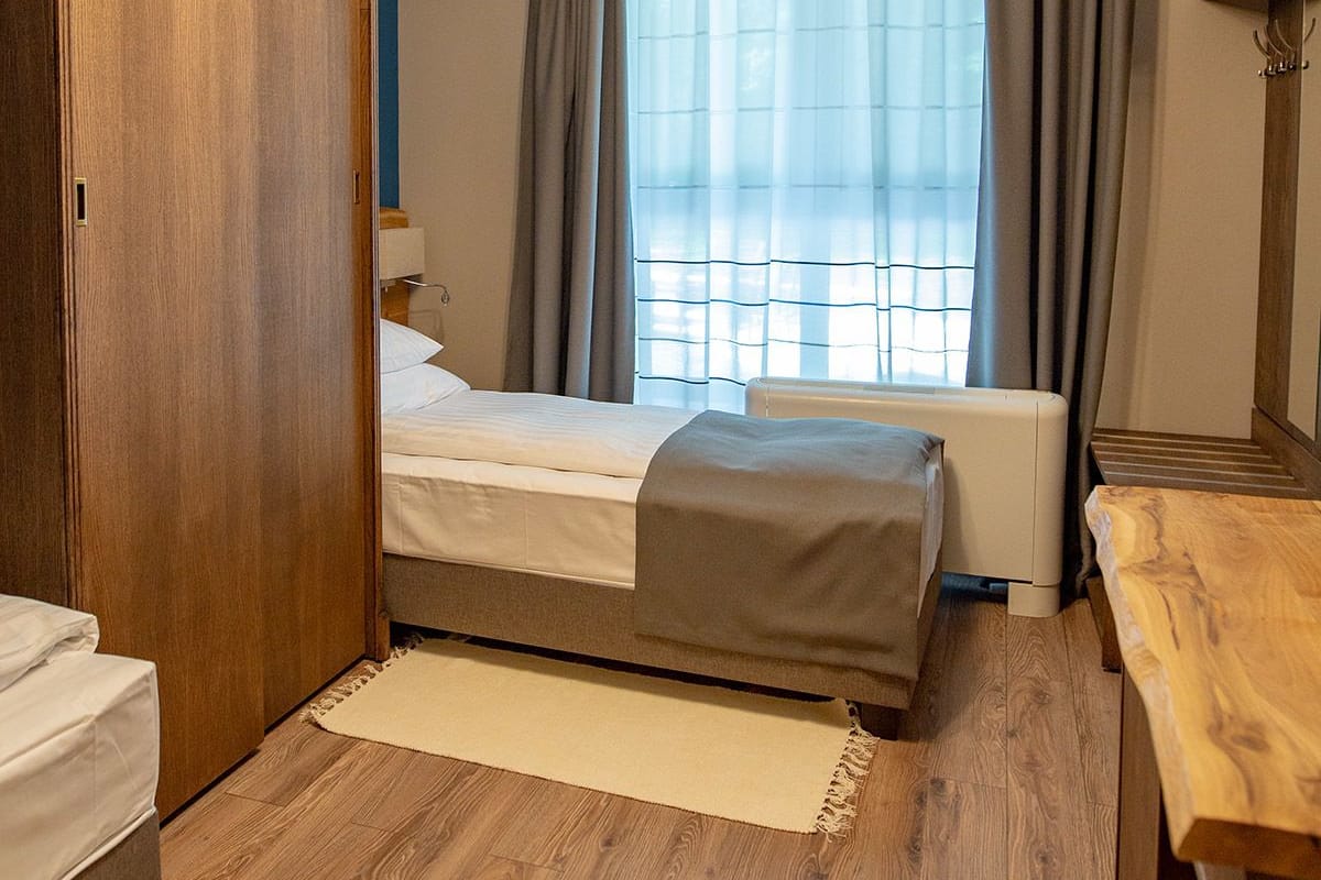 Een slaapkamer met een bed, een houten kledingkast, een nachtkastje, een raam met gordijnen en een radiator.
