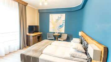 A hotel room with a bed, a desk with a TV, two chairs, a small table with glasses, and a large abstract painting on the wall.