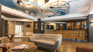 Um lobby moderno com área de bar, poltrona estofada e mesa de madeira redonda com revistas.