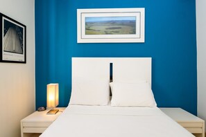 Un lit bien fait, avec du linge de lit blanc et des oreillers, une lampe de chevet et une photographie encadrée représentant un paysage accrochée au mur.