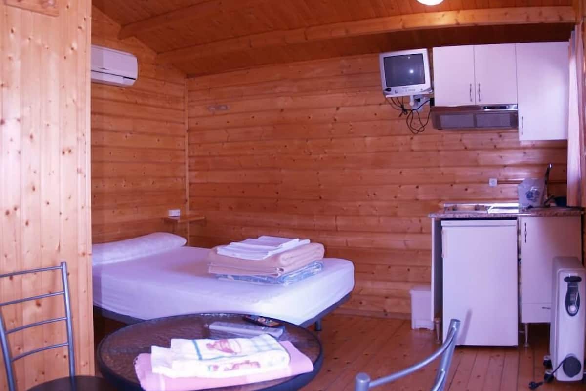 A small wooden cabin with a single bed, a small table with a chair, a kitchenette, and a television.