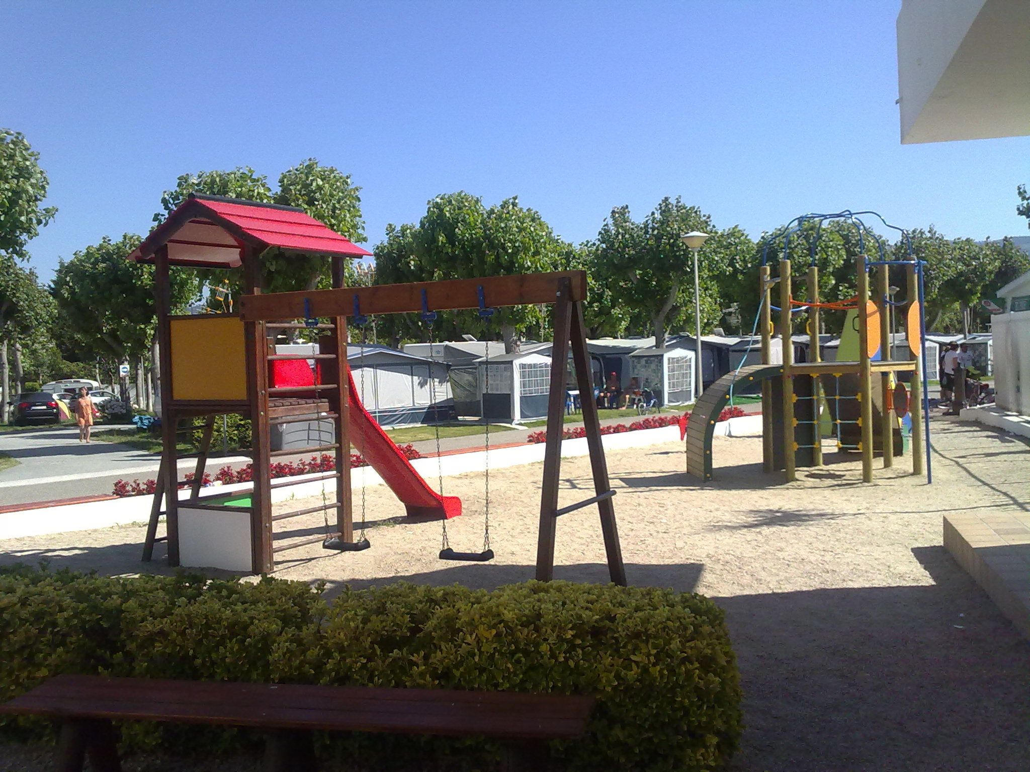 A playground with a red slide and swings, surrounded by greenery and a road with parked cars.