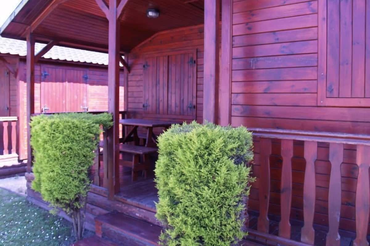 A wooden cabin with a porch, a table, and two neatly trimmed bushes.