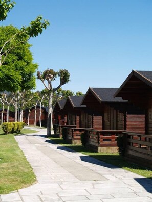 A row of wooden cabins with a paved walkway and trees. - Camping Playa América (Nigran)