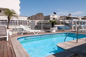 Ein Pool auf dem Dach mit weißen Liegestühlen, einer Palme und Blick auf einen felsigen Hügel.