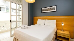 Una cama bien hecha con sábanas blancas, cabecera de madera, lámpara de noche y un cuadro enmarcado en la pared.