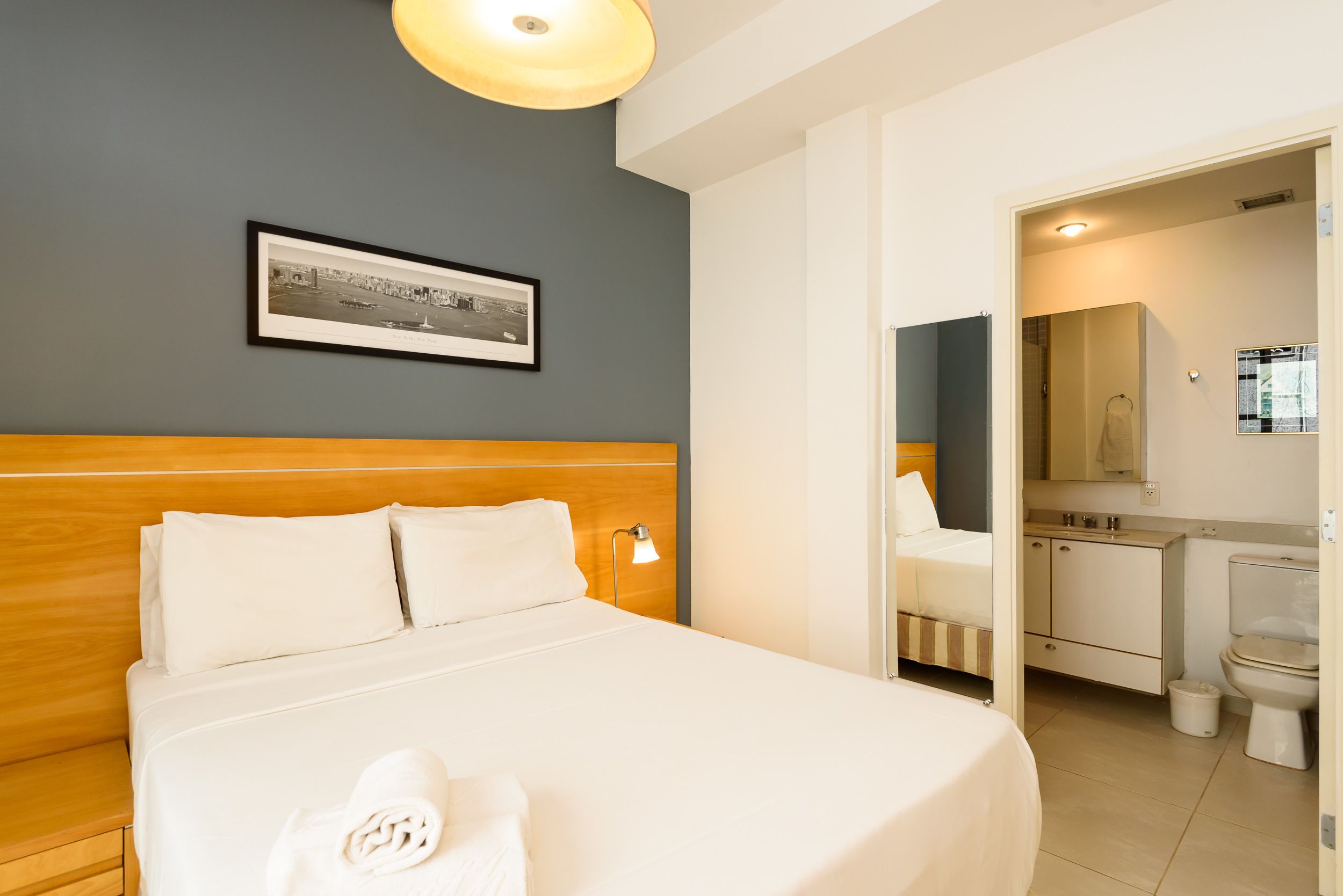 Une chambre d’hôtel avec une tête de lit en bois, un lit soigneusement fait, un tableau encadré accroché au mur et une salle de bain visible à travers une porte ouverte.