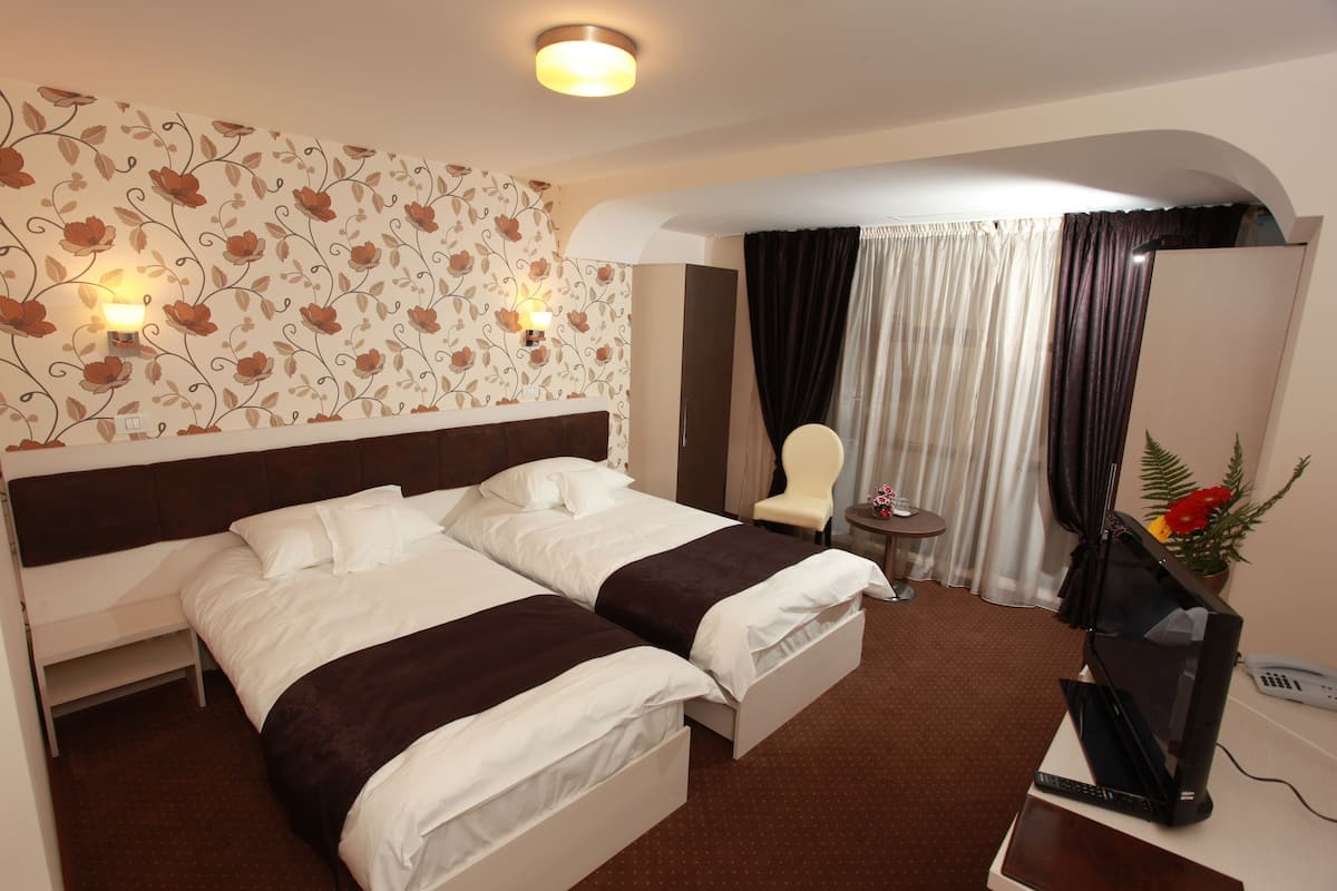 A hotel room with two beds, a desk with a computer, a chair, a small table with a lamp, and a decorative plant.