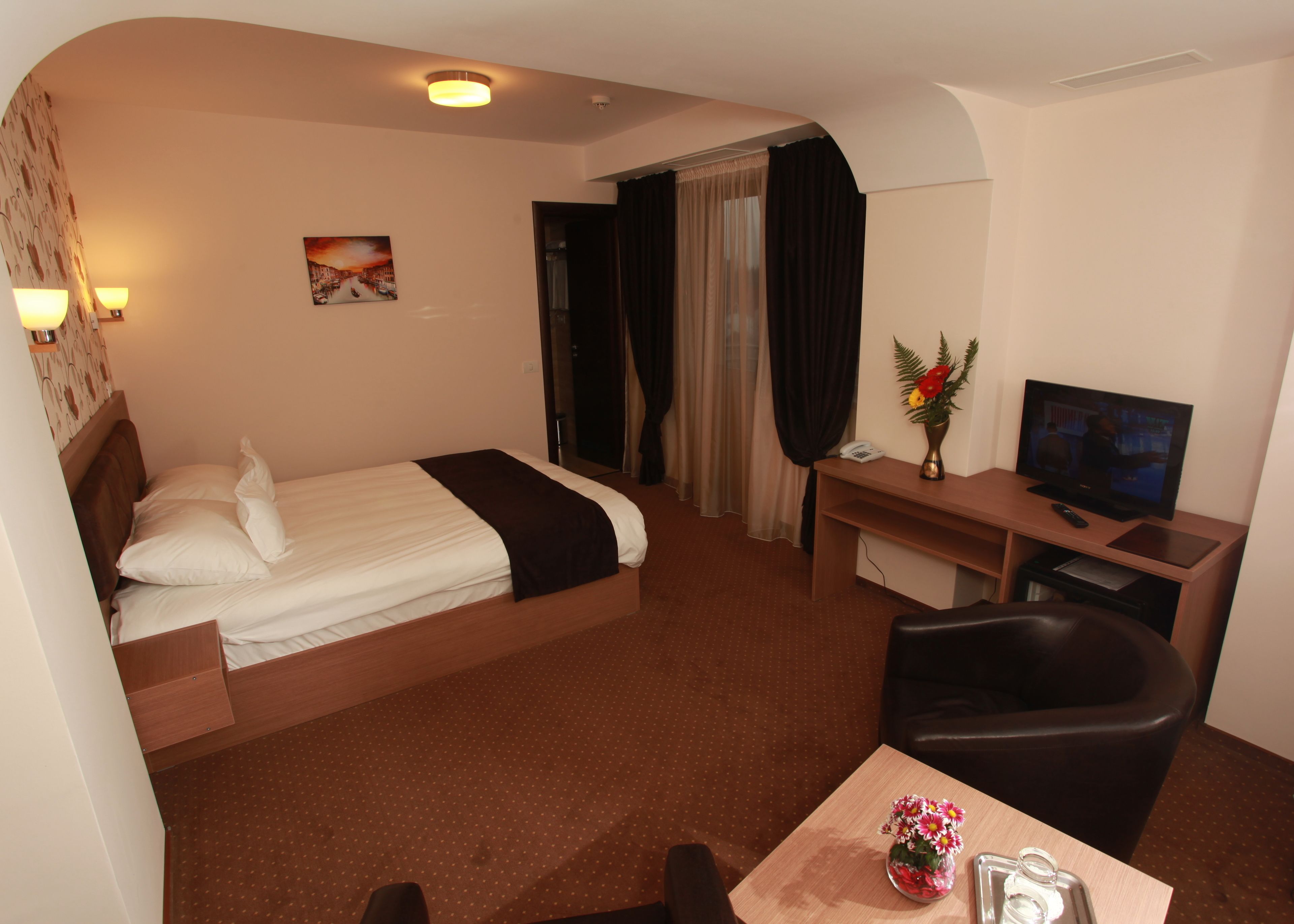 Una habitación de hotel con una cama, un escritorio, un televisor, una silla y una mesita con un florero.