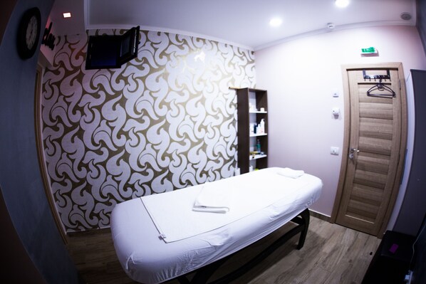 Una habitación con cama, televisor de pared, puerta de madera y estante con artículos de aseo.