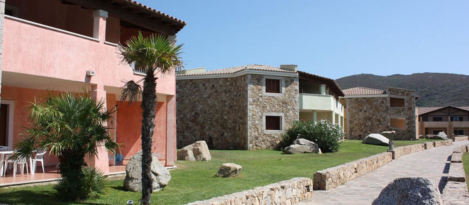 A resort with stone buildings, a paved walkway, and a grassy area with palm trees.