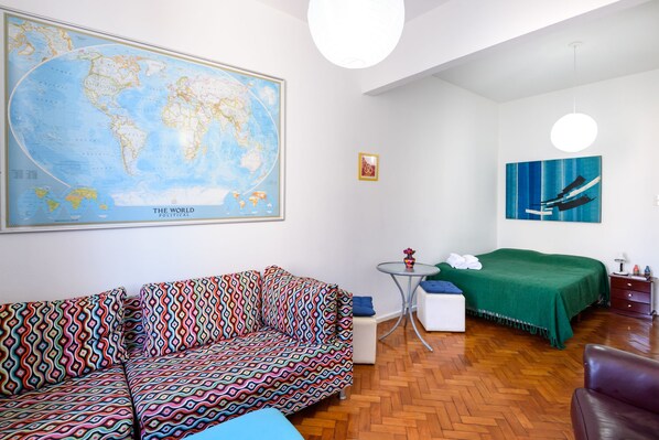 A room with a large world map, a colorful sofa, a bed with a green blanket, a small round table with a vase, and a hanging light fixture. - Omar do Rio - SL906 (Rio de Janeiro)