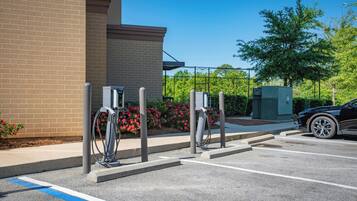 A parking area with electric vehicle charging stations, a brick building, and a tree in the background.