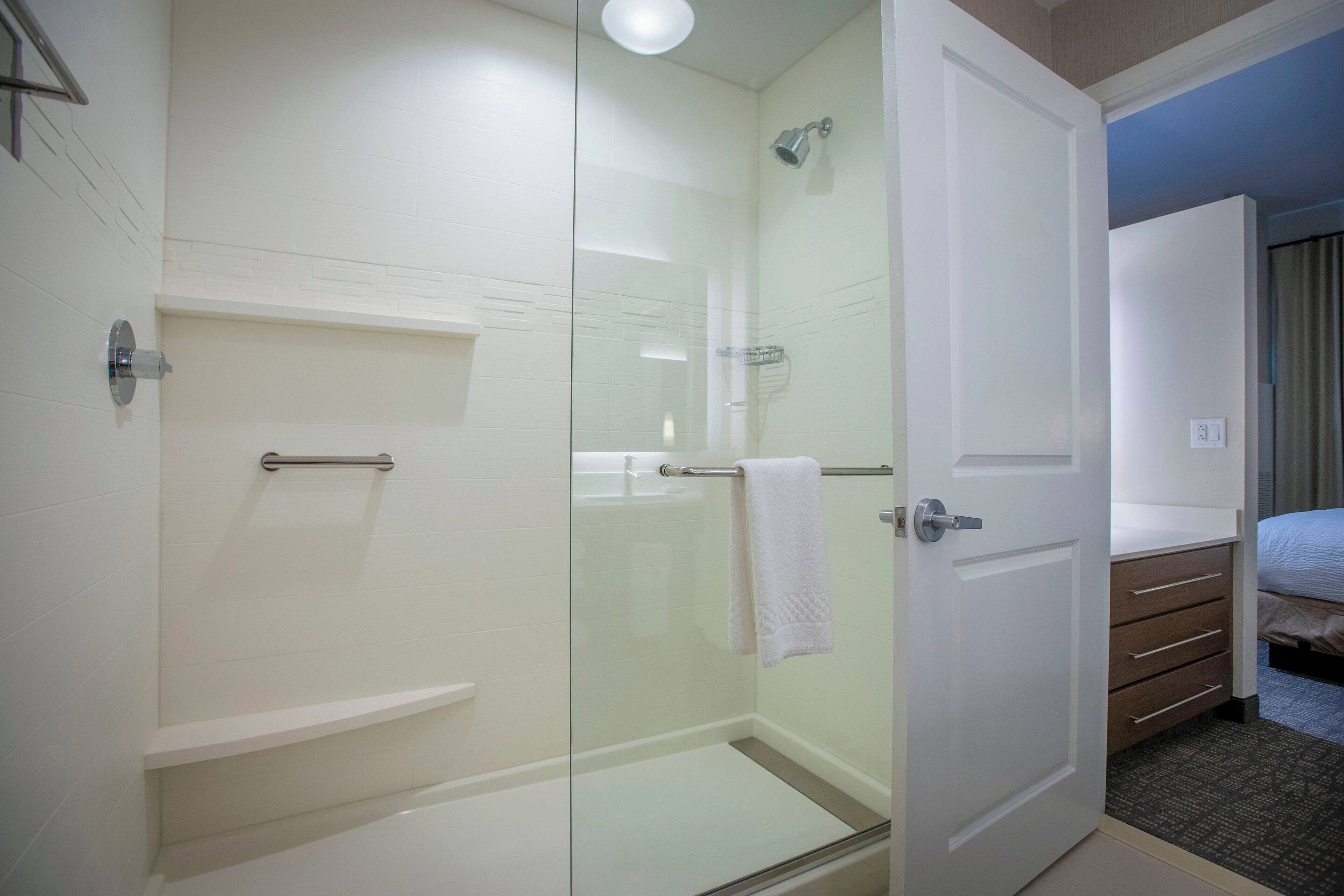 Un baño de hotel con ducha envuelta en cristal, una toalla blanca colgada en la puerta de la ducha y una cama de madera en la habitación contigua.