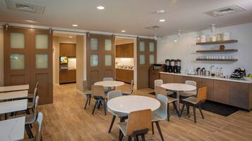 A modern dining area with round tables and chairs, wooden flooring, and a kitchen area with wooden cabinets and shelves.