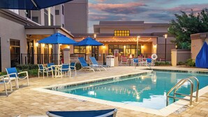 Uma área de piscina com espreguiçadeiras e guarda-sóis, uma piscina com placa indicando a profundidade e um edifício iluminado ao fundo.
