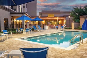 Un espace piscine avec des chaises longues et des parasols, une piscine avec un panneau indiquant la profondeur, et un bâtiment illuminé en arrière-plan.