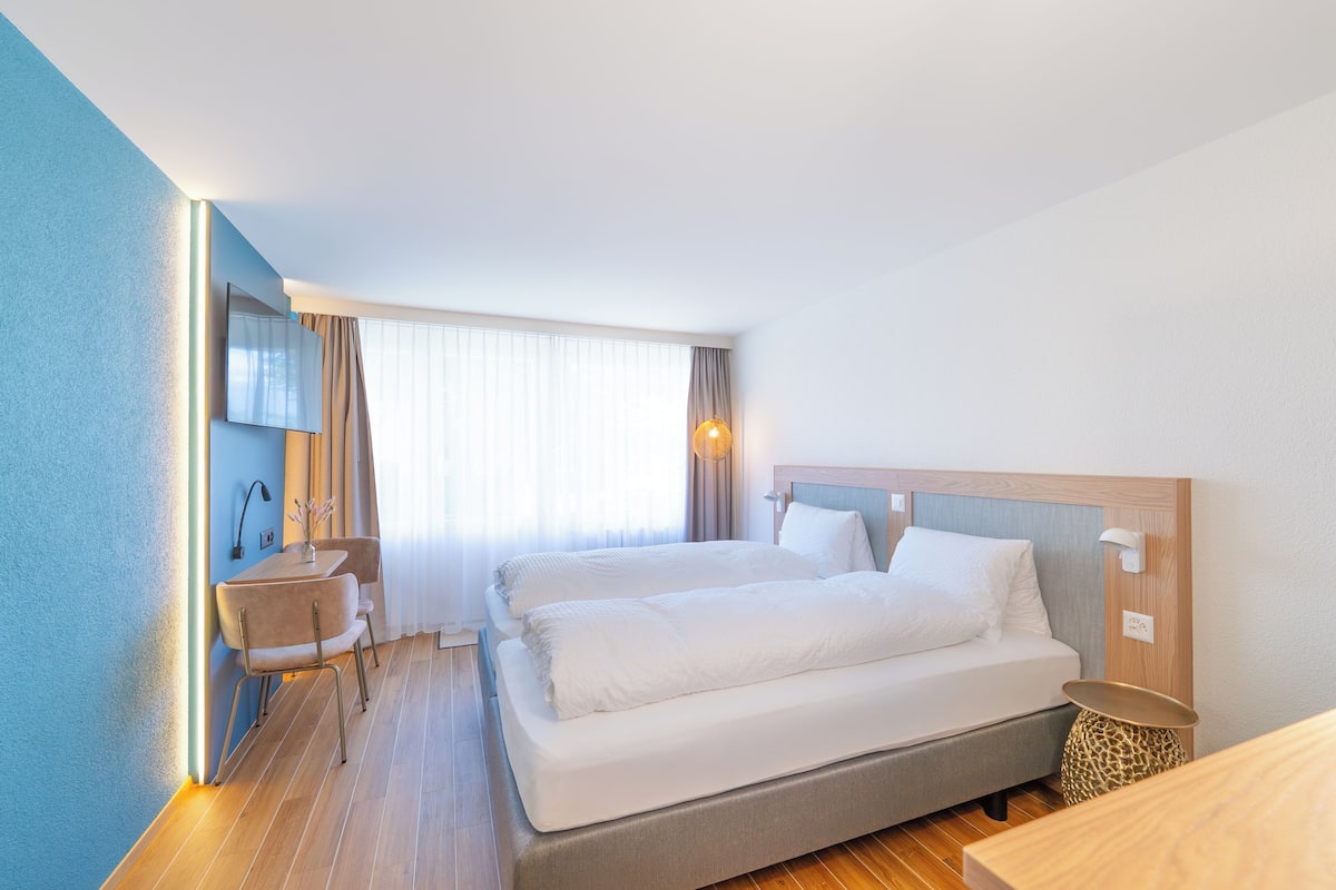 A hotel room with a bed, a desk, a chair, a lamp, and a window with curtains.