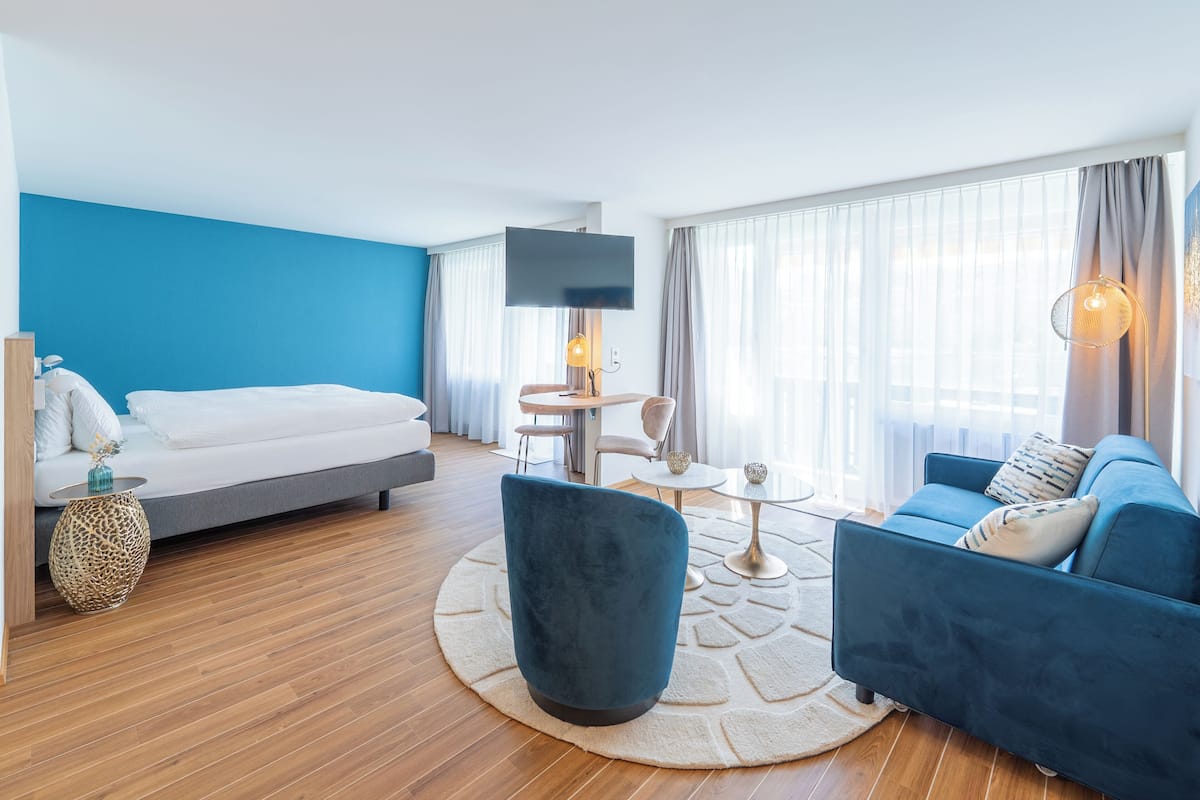A modern hotel room with a bed, blue armchair, sofa, and a TV.