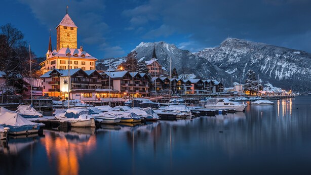 Un bâtiment historique doté d’une tour, entouré de montagnes enneigées et bordé d’un lac paisible où flottent des bateaux.