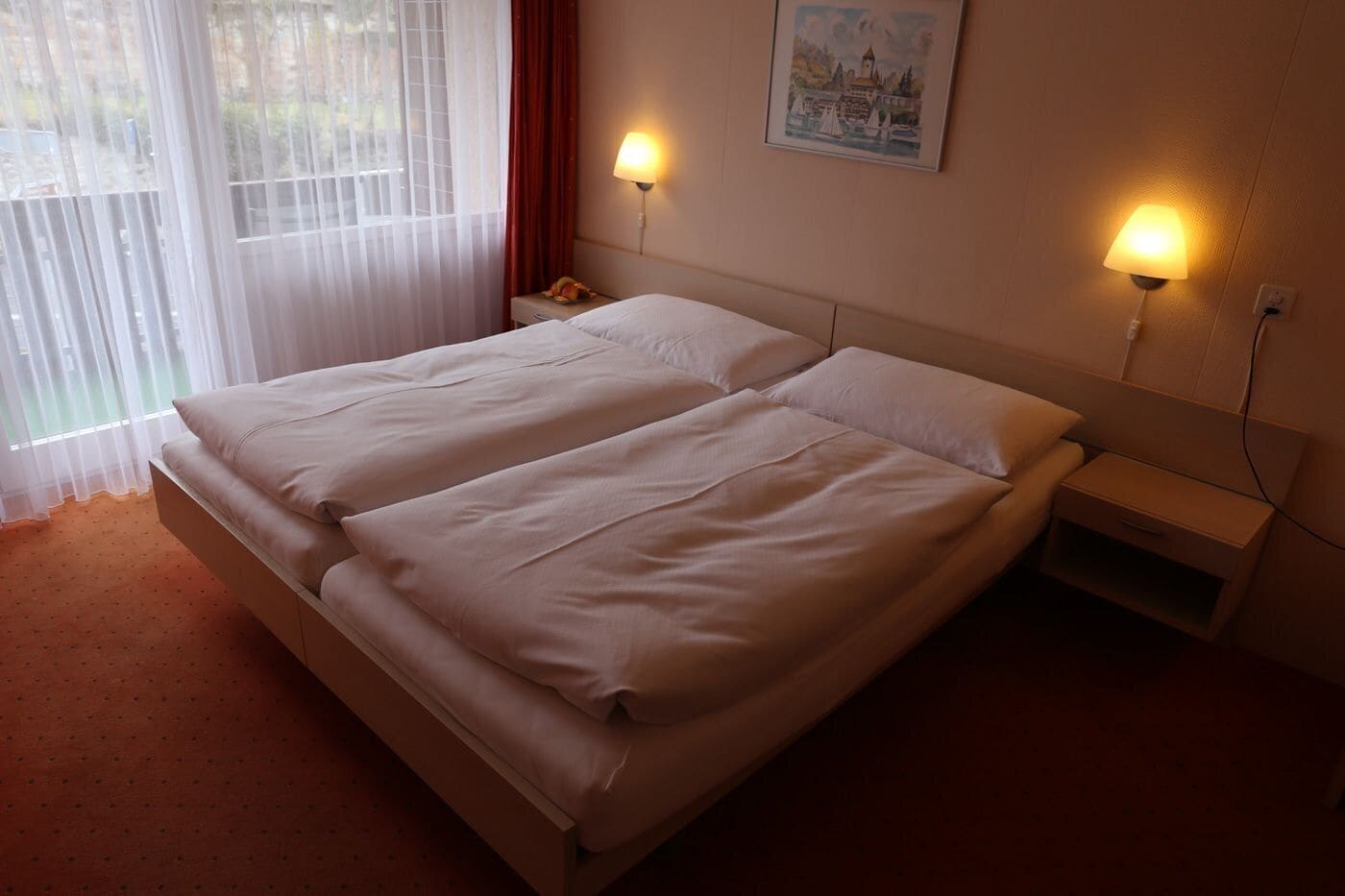 Un lit double avec deux tables de chevet, un tableau au mur et une fenêtre avec des rideaux légers.