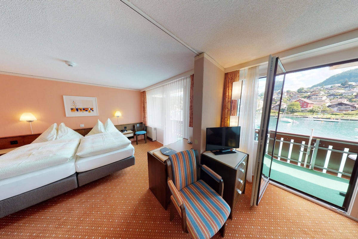 A hotel room with a bed, a desk with a chair, a television, and a large window offering a view of a body of water and buildings.