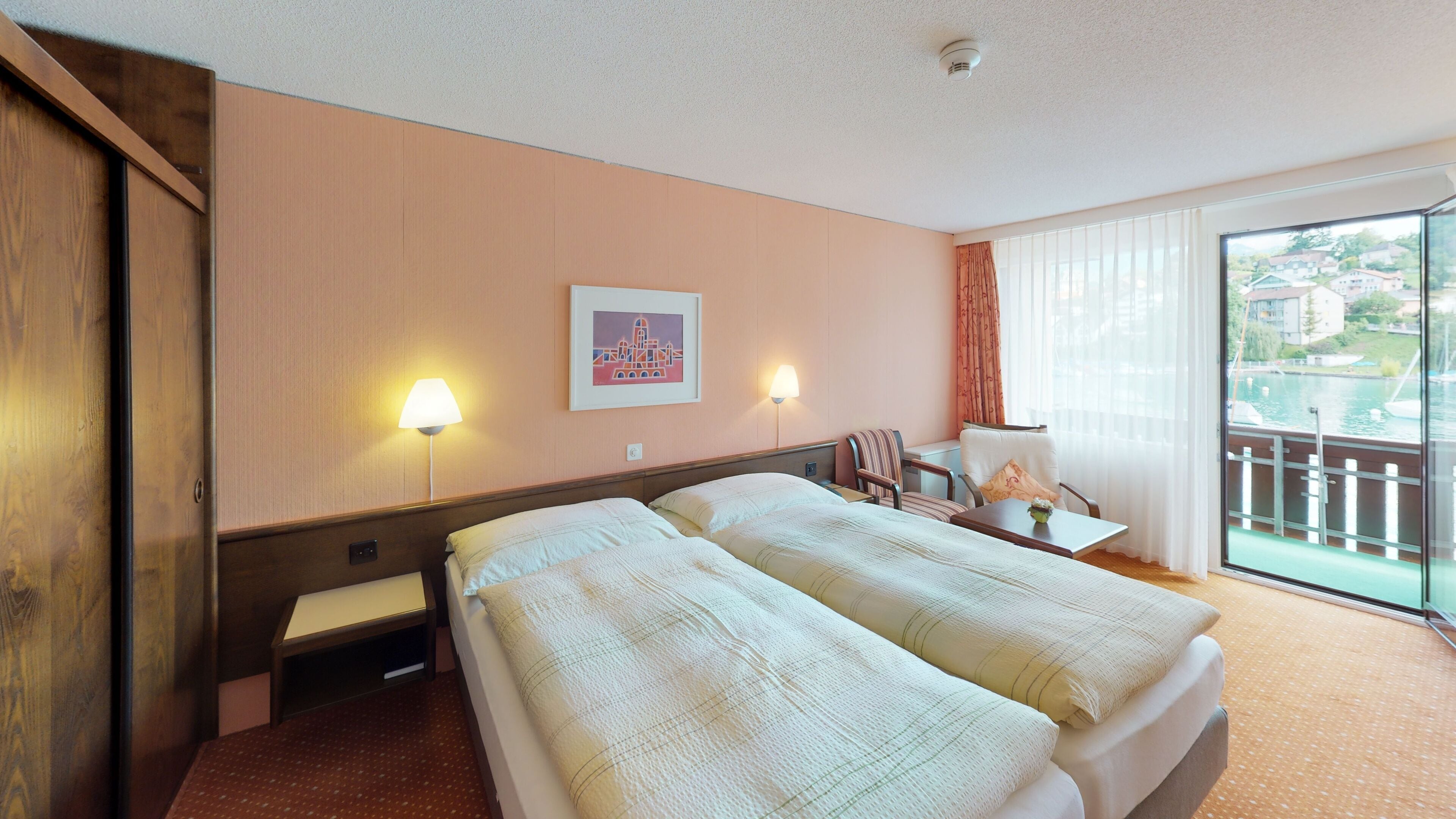 A hotel room with two beds, a desk, a chair, a lamp, and a window with a view of a body of water and buildings.