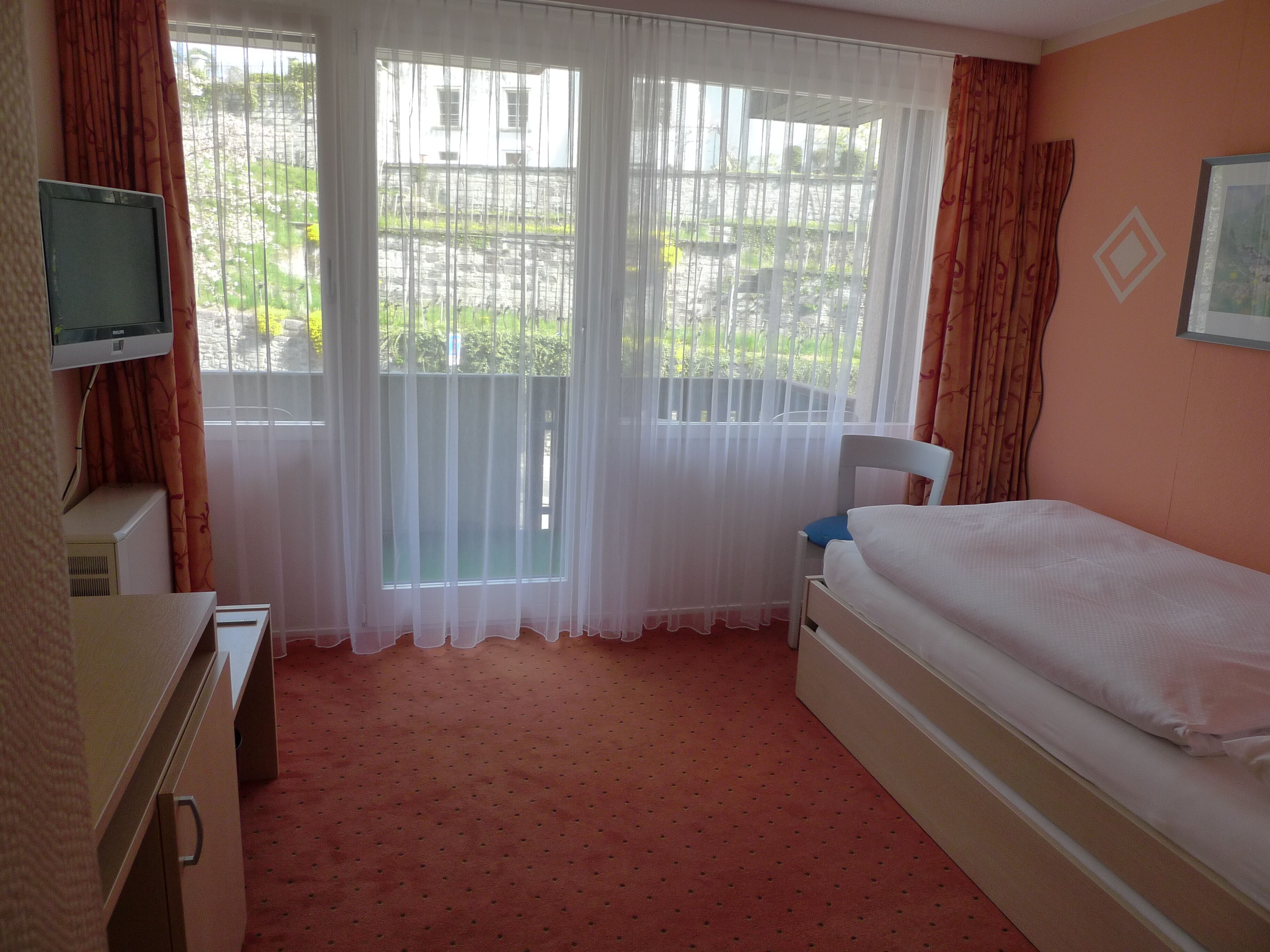 A hotel room with a bed, a desk with a television, and a window with curtains.