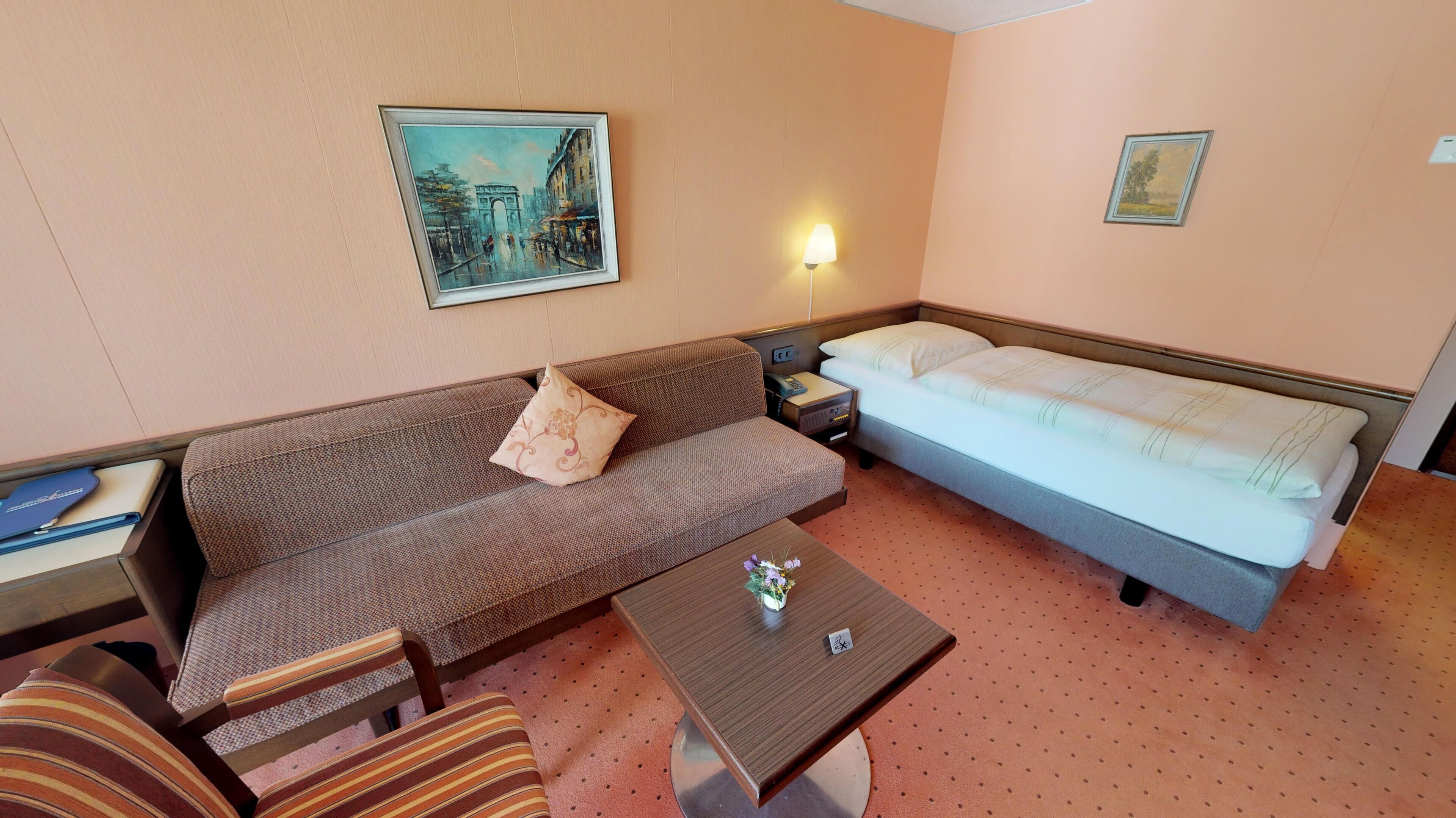 A hotel room with a sofa, a single bed, a small table with a vase, a nightstand, and a lamp.