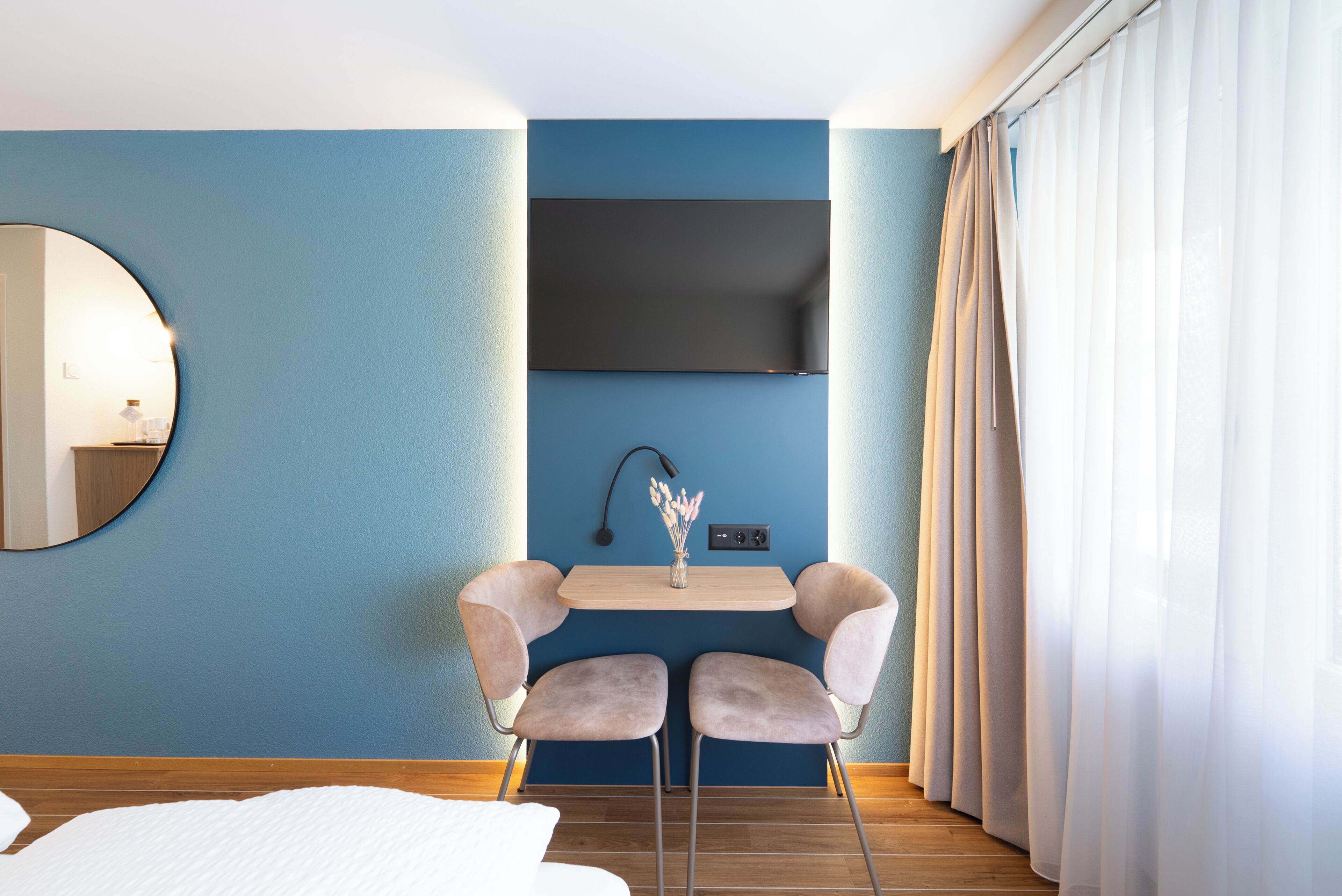 A hotel room with a blue wall, a flat-screen TV mounted above a small table with two chairs, a round mirror, and a window with white curtains.